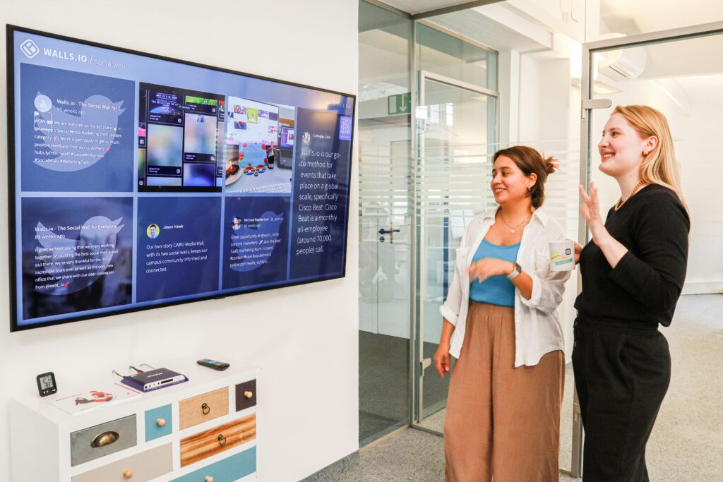 Two women in an office environment interact with a large digital display showcasing social media content.